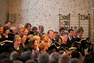 Kirchenchor singt, hält Gesangbücher, Liedtafel im Hintergrund.