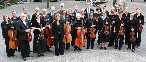 Orchestergruppe, meist Erwachsene, halten Streichinstrumente im Freien.