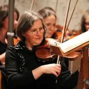 Frau mit Brille spielt Geige, Noten vom Ständer lesend.