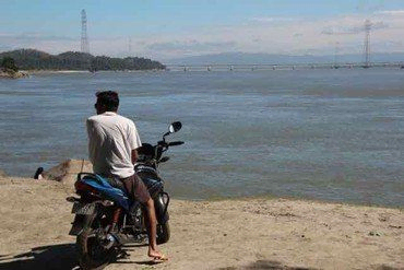 Mann auf Motorrad blickt auf weiten Fluss und ferne Brücke.